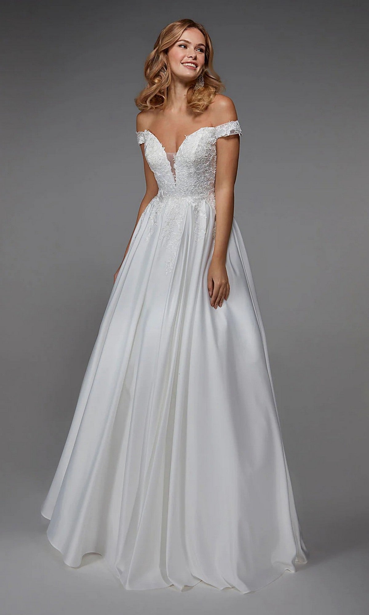 A woman wearing the Alyce 7034 long diamond white dress, featuring an off-the-shoulder floral embroidered bodice and full satin skirt, stands against a plain gray background.